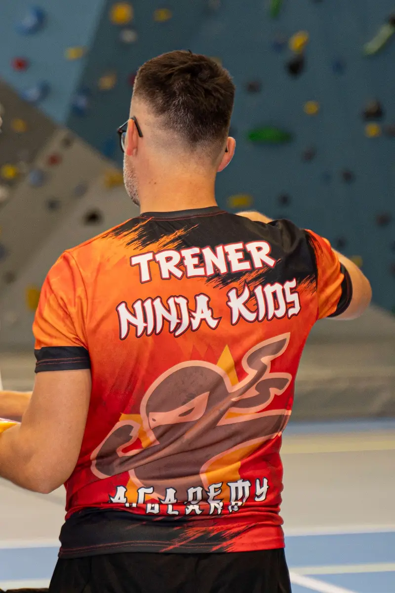 Trener Ninja Kids Academy stojący odwrócony do kamery. Posiada koszulkę trenera Ninja Kids Academy z logo
